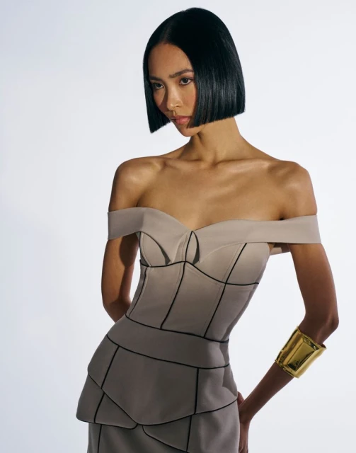 Woman with an Asian appearance poses in a studio. She is wearing an off-the-shoulder dress and a wide gold cuff bracelet on her arm.