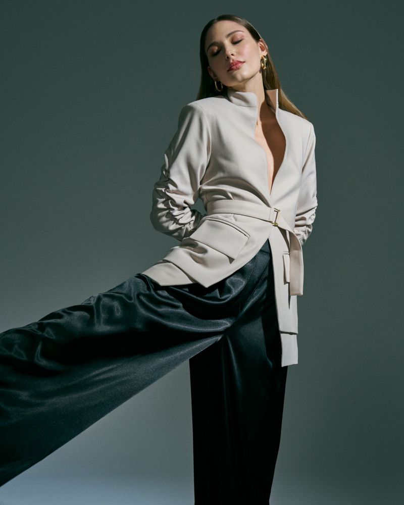 Woman poses in a beige blazer and black wide-leg pants against a gray background, eyes closed, gold hoop earrings, neutral colors.