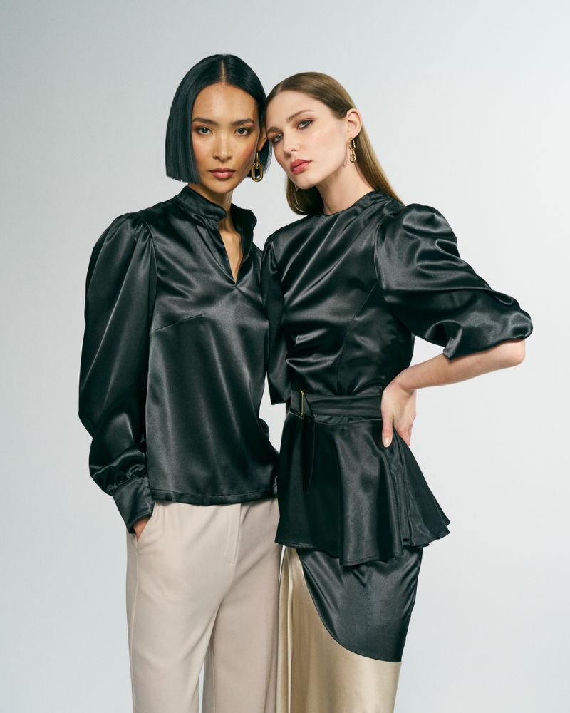 Two women in fashionable satin blouses and stylish pants pose in a studio, radiating elegance and sophistication in a fashion editorial setting.