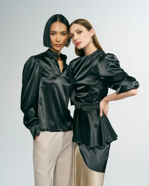 Two women in fashionable satin blouses and stylish pants pose in a studio, radiating elegance and sophistication in a fashion editorial setting.