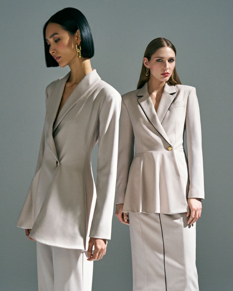 Two women in fashionable blazers stand in a photo studio. The models wear neutral-toned blazers with minimalistic gold earrings.
