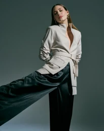 Model in cream belted jacket and dark satin trousers posing against a grey studio backdrop