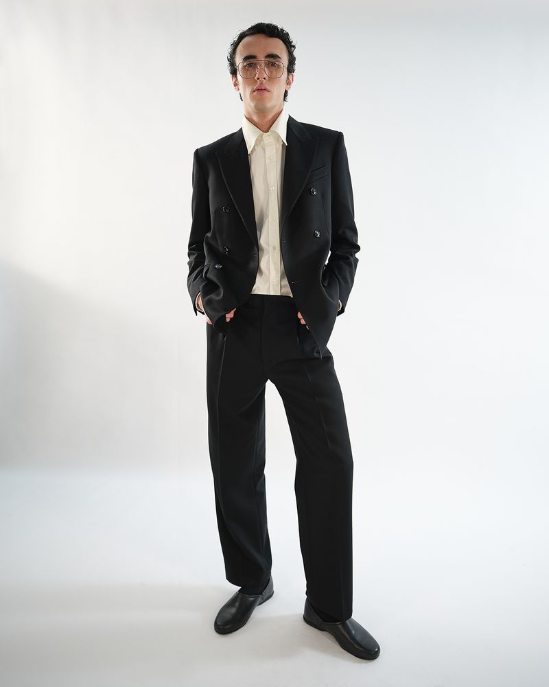 A man wearing a black double-breasted suit and a light-colored dress shirt with glasses, standing against a plain white background.