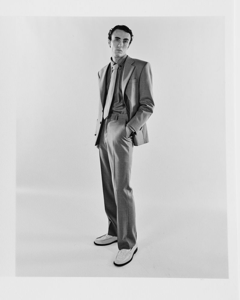 Full-length portrait of a man in a suit with tie, posing in a photo studio with a neutral expression. He wears shoes.