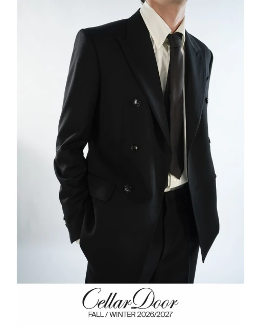 Man in a black double-breasted suit and black tie, standing against a white backdrop, showcasing formal menswear fashion.