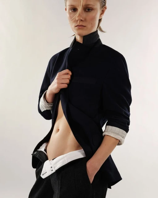Blonde woman in fashion pose, wearing a navy jacket and dark pants. She stands against a light gray background in a studio shot.