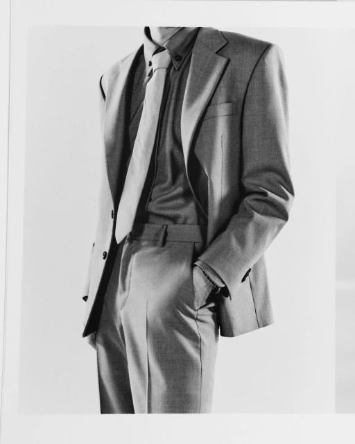 Monochromatic studio shot of a torso in a grey suit, buttoned shirt, and matching tie, hand casually tucked in pocket on a light background.