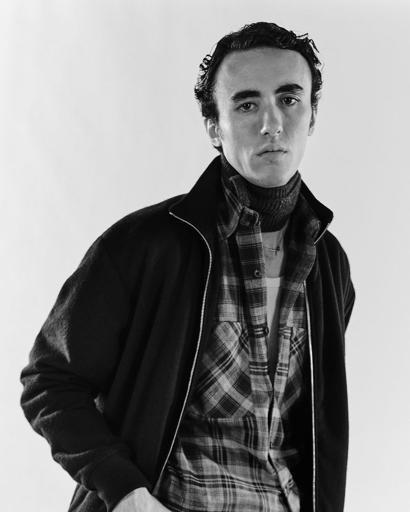 Monochrome portrait of a young adult man wearing a turtleneck, flannel shirt, and jacket, with a neutral facial expression and studio lighting.