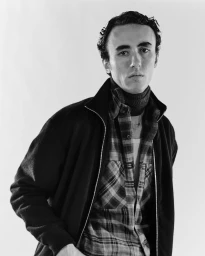 Black and white portrait of a man wearing an open black zip-up jacket over a plaid shirt and turtleneck against a plain white background.