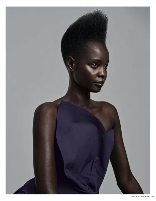 Portrait of a Black woman with a distinctive updo, wearing an asymmetrical navy dress, against a plain grey background in a studio setting.