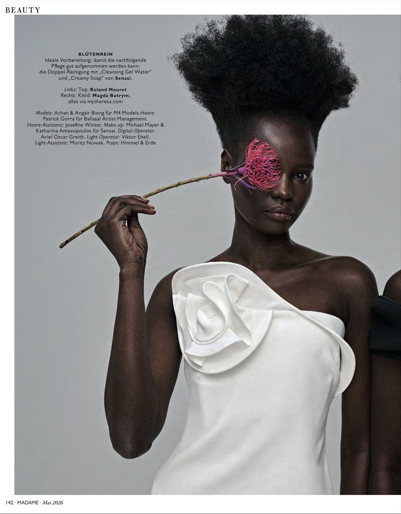 Beauty editorial shot of Black model with Afro holding pink flower in front of eye, wearing white, one-shoulder dress in a photo studio.