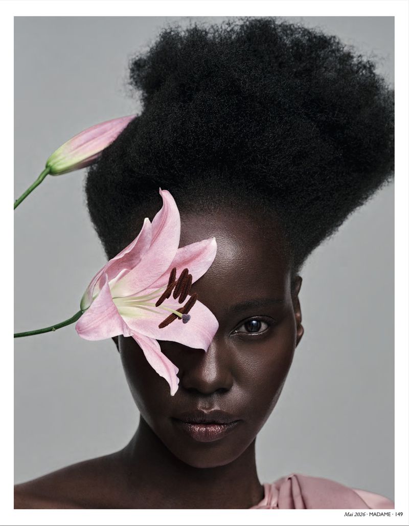 Close-up portrait of a dark-skinned woman with a large afro hairstyle, adorned with a pink lily covering one eye against a grey backdrop.