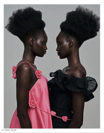 Two dark-skinned women face each other wearing pink and black dresses with floral accents, set against a neutral backdrop in a studio shot.
