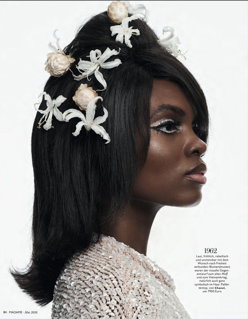 Profile portrait of a woman with 1960s hairstyle adorned with white flowers, wearing a shimmering top against a white background.