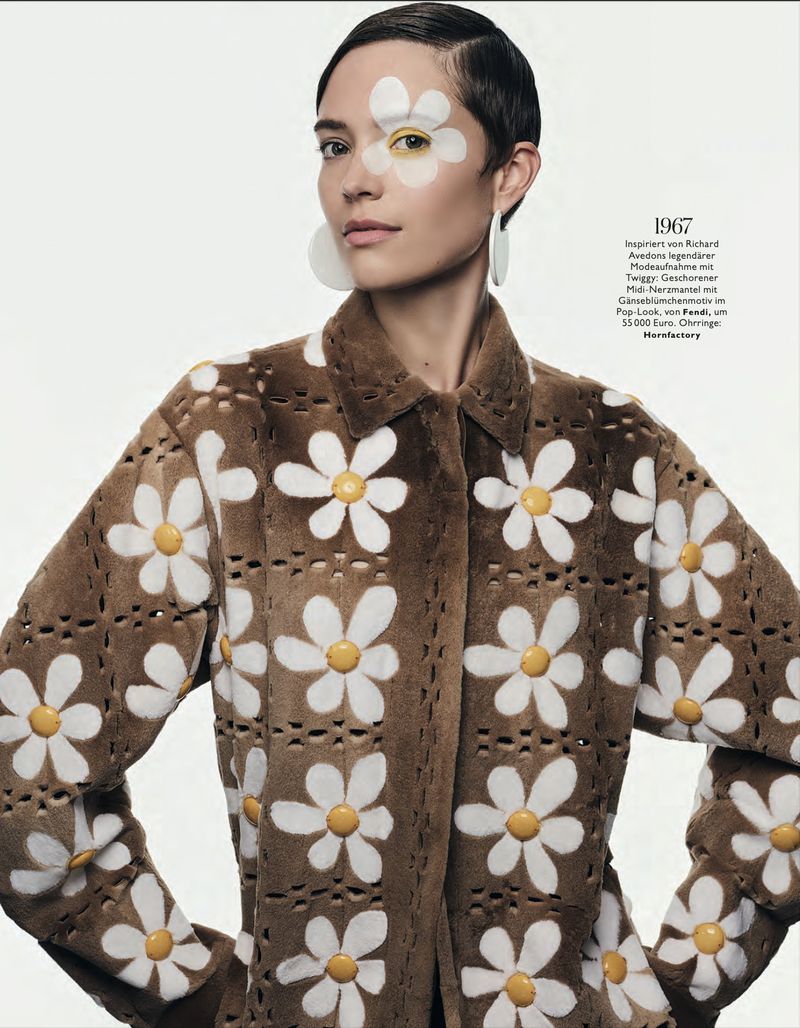 Fashion shot of a woman in a daisy-patterned coat with matching eye makeup and large earrings posing in front of a white background.