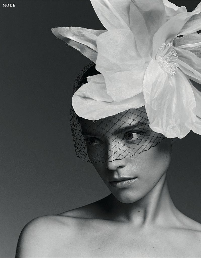 Monochrome portrait of a woman wearing a large white floral hat with a black veil, gazing intently at the viewer in a studio setting.
