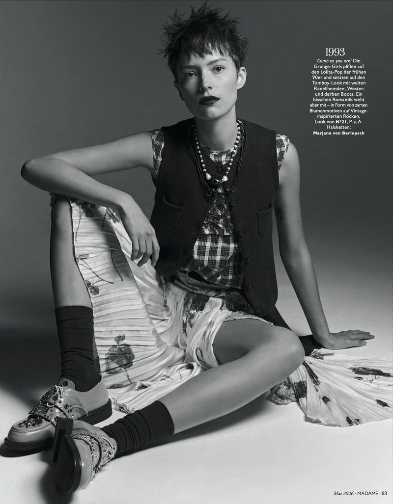Monochrome studio shot of a young adult woman with short hair, wearing a vest, plaid shirt, pleated skirt, socks, and shoes, seated.