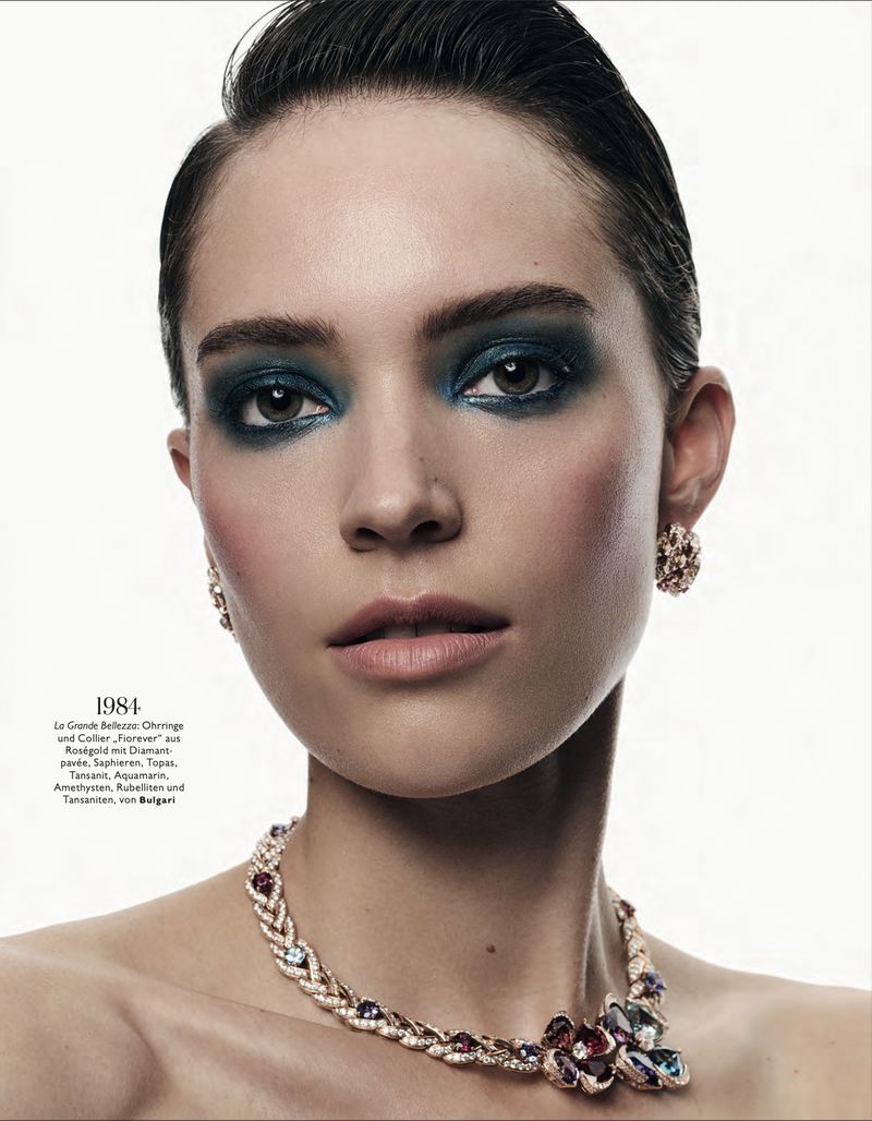 Close-up portrait of a woman with slicked-back hair, bold dark teal eye makeup, and an ornate gemstone necklace against a white backdrop.