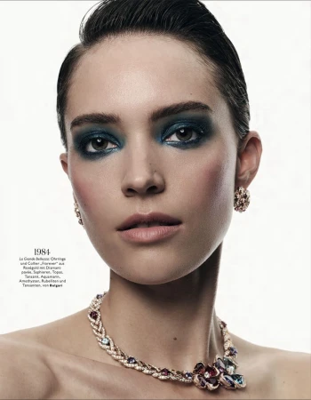 Close-up portrait of a woman with slicked-back hair, bold dark teal eye makeup, and an ornate gemstone necklace against a white backdrop.