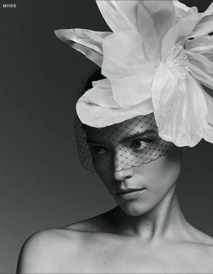 Black and white portrait of a woman wearing a large white fabric flower headpiece with a mesh face veil against a neutral studio backdrop.