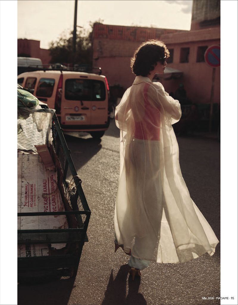 Woman in a sheer white trench coat walks on a sunny street in an urban setting, with a yellow van and buildings in the background.
