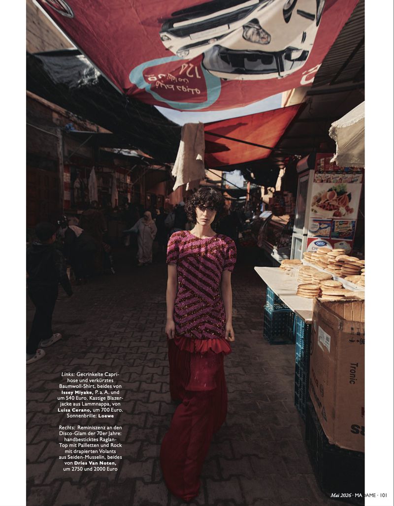 A woman with curly hair models a pink and red dress with striped accents in a bustling outdoor market with colorful awnings.