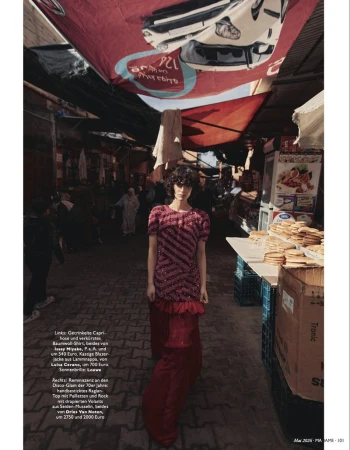 A woman with curly hair models a pink and red dress with striped accents in a bustling outdoor market with colorful awnings.