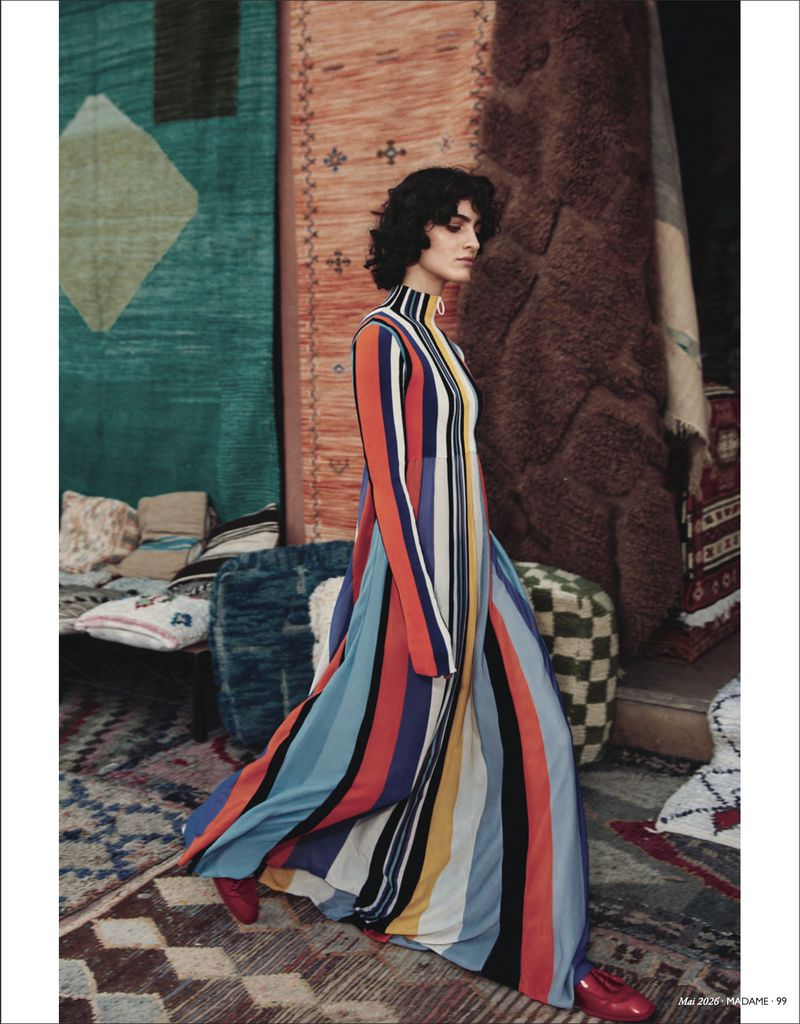 Fashion editorial shot of a woman in a striped long-sleeve dress, walking across a rug-covered floor with patterned textiles in the background.