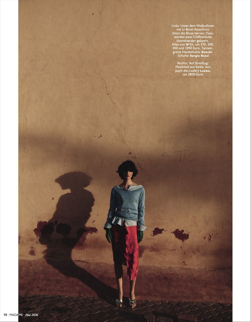 Fashion editorial shot of a woman in a blue top and red skirt posing against a textured tan wall with strong shadows, creating a dramatic scene.
