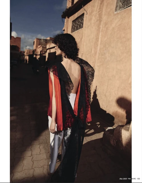 Woman in red top and lace-trimmed dress poses in an alley with peach-colored buildings and shadows cast by the sun. Fashion editorial.