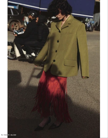 A model wearing a structured olive green blazer and a long red fringed skirt stands on a paved street with a scooter in the background.