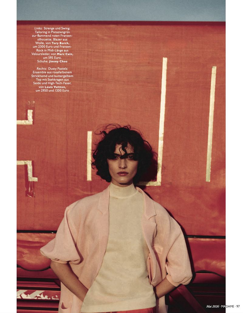 Fashion editorial shot of a woman with dark curly hair in a beige turtleneck and light pink jacket in front of an orange backdrop.