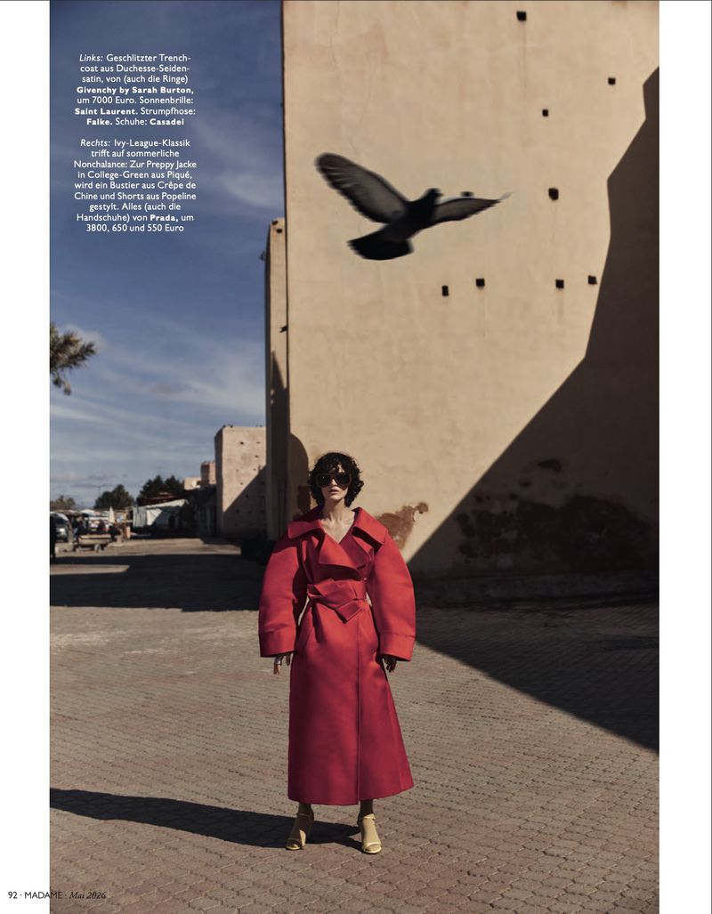 A woman in a red coat stands in an outdoor plaza with a large building in the background and a bird flying overhead.