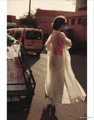 Model wearing sheer white coat and trousers walking down an urban street past a parked van and cart