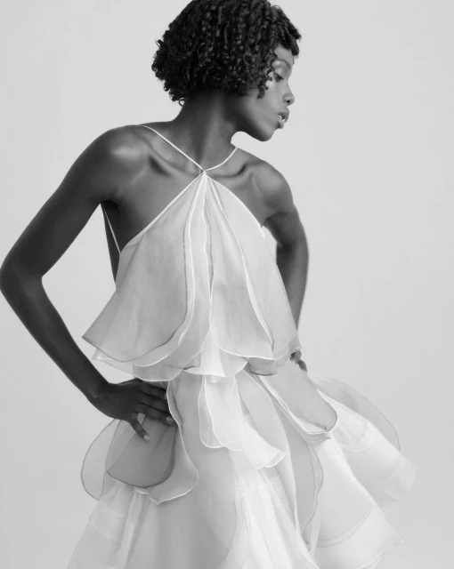 Black female model in a layered white halter dress poses in a studio. The dress has a petal-like pattern. Black and white portrait.