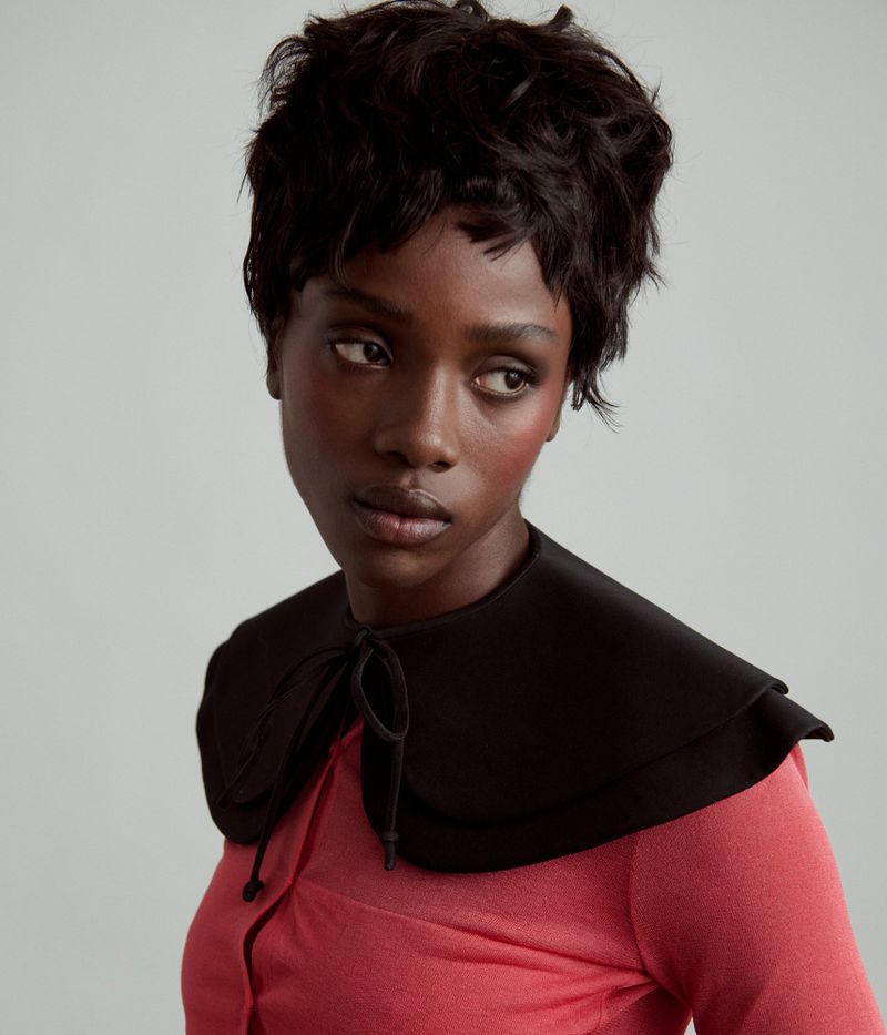 A dark-skinned woman with a short, wavy hairstyle wears a black Peter Pan collar and a pink top in a studio portrait.