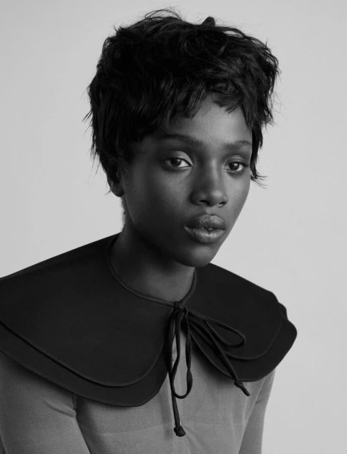 Monochrome portrait of a young Black female model with short hair wearing a large black collar with tie strings against a plain studio backdrop.