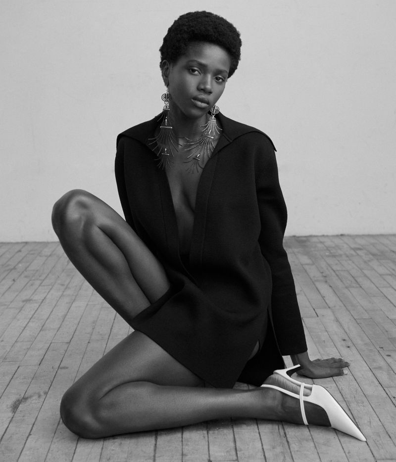 Black and white studio shot of an adult African American woman wearing a dark jacket and light-colored high heels, posing on a wood floor.