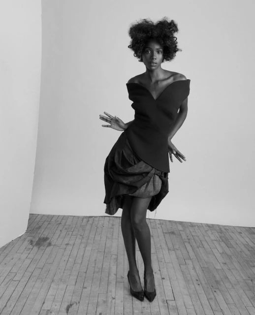 Black and white portrait of a black female fashion model in an off-the-shoulder top, skirt, and heels, posing in a photo studio.