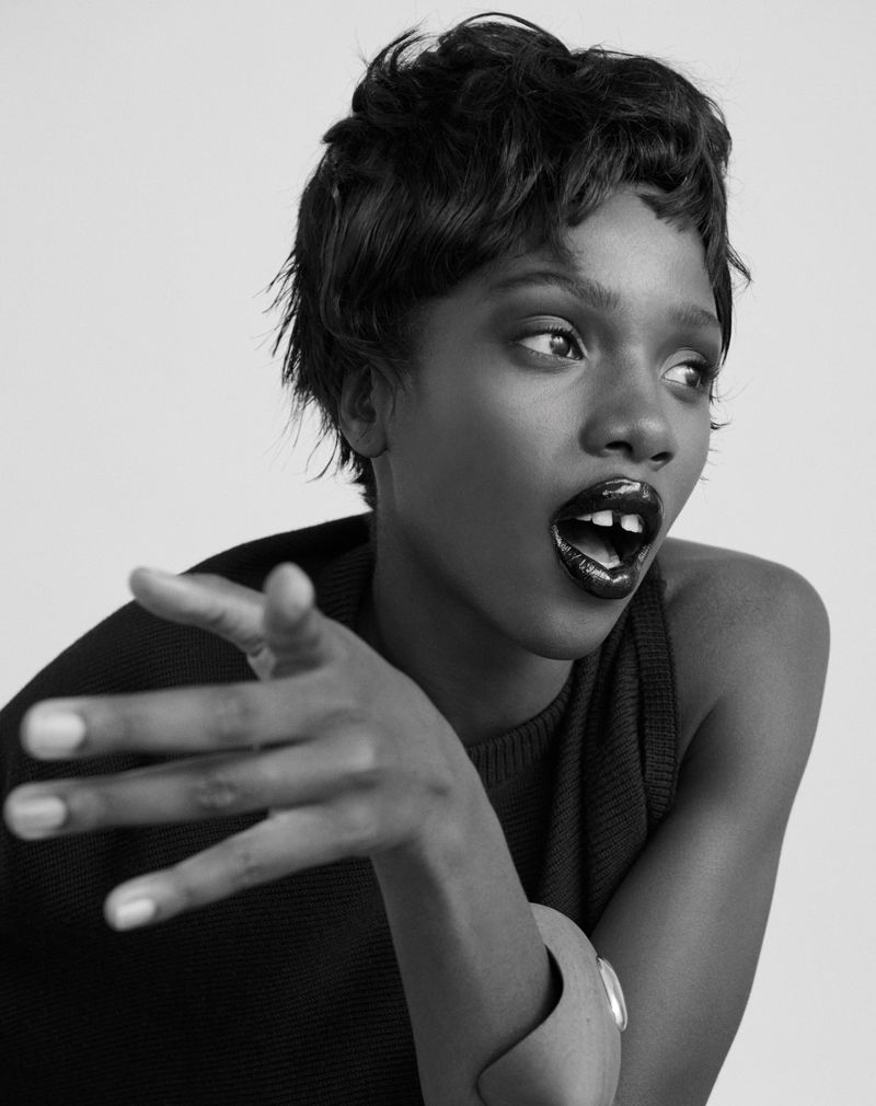 Black and white portrait of a Black female model with short hair, dark lipstick, and a bracelet gesturing toward the right of the frame.