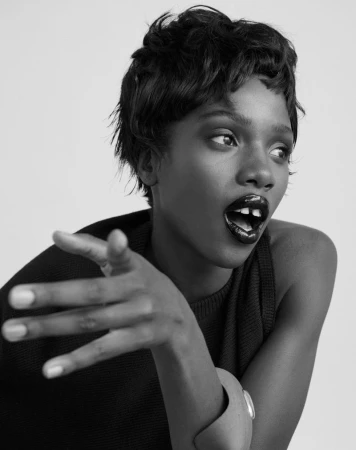 Black and white portrait of a Black female model with short hair, dark lipstick, and a bracelet gesturing toward the right of the frame.