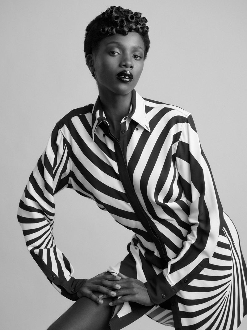 Black and white portrait of a striking Black female model in a boldly striped shirt dress, gazing directly at the viewer in a photo studio.