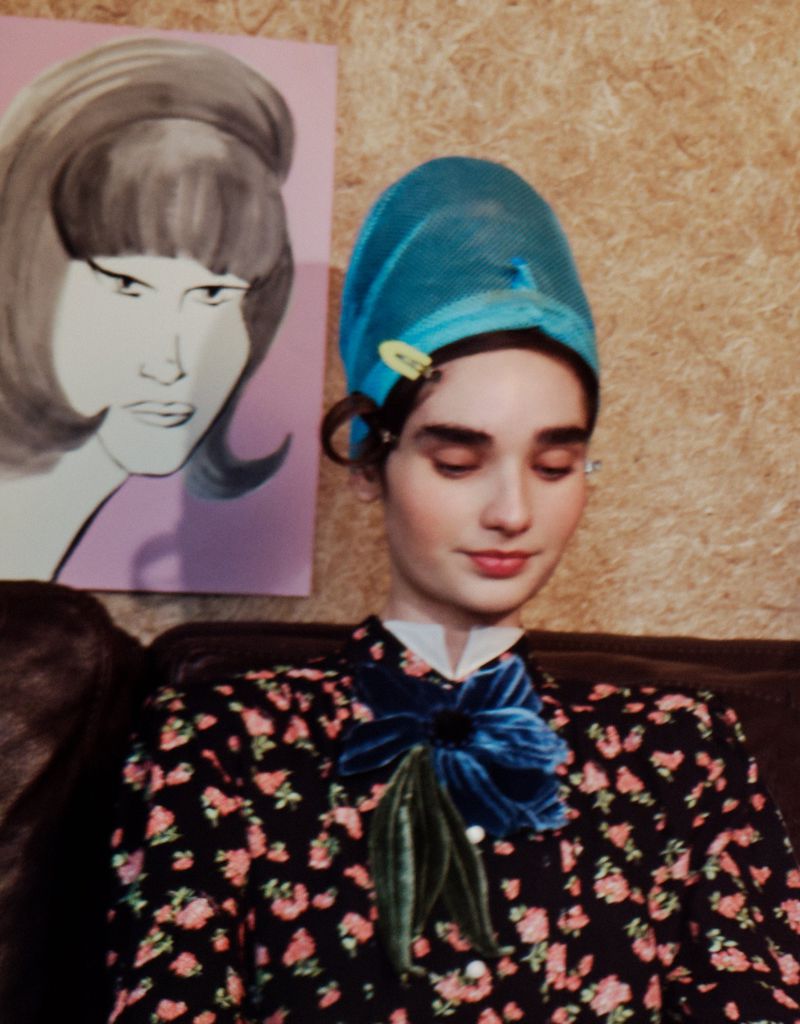 Woman in floral dress and blue hair wrap poses indoors with portrait art nearby. Portrait features mid-century hairstyle.