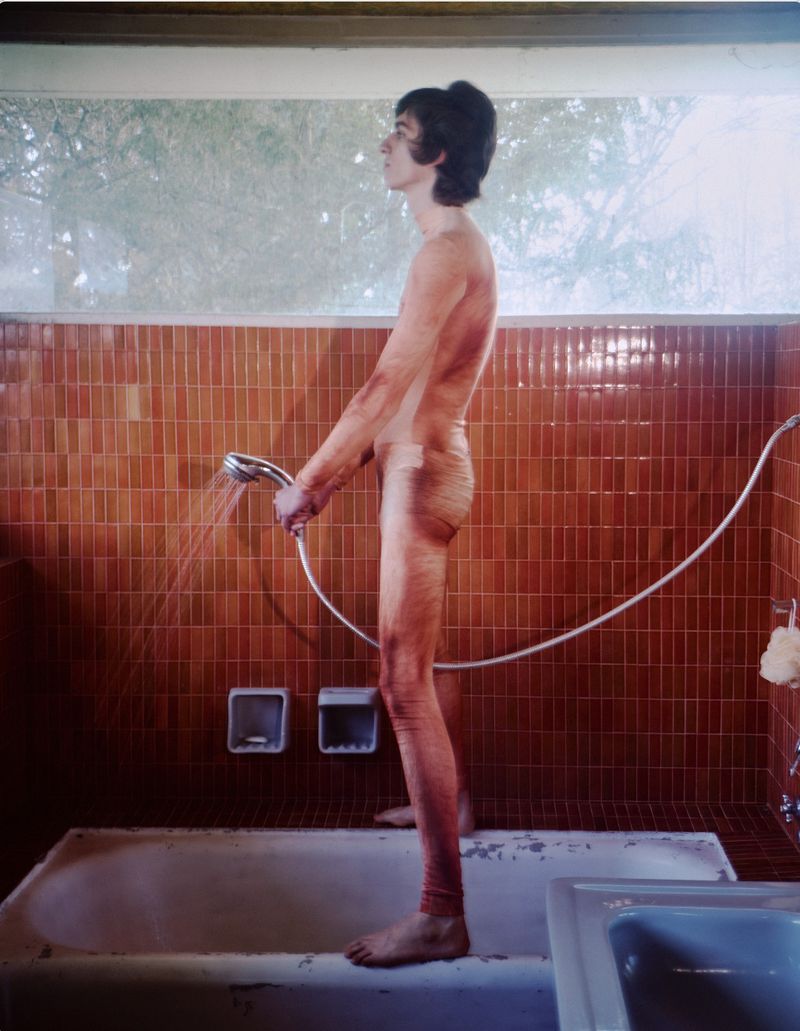 Nude adult male standing in a bathroom, holding a showerhead with water spraying. Brown tiled walls and a bathtub are visible.