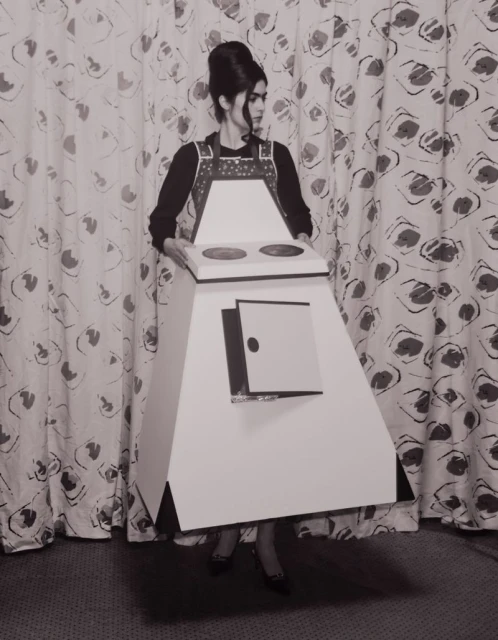 Woman in stove costume standing against patterned backdrop. Black and white photograph of retro fashion and creative portrait.