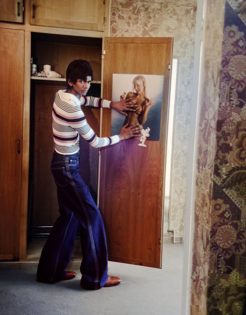 Man in striped shirt and bell-bottom jeans poses with a portrait of a woman attached to a wooden door in an indoor setting.
