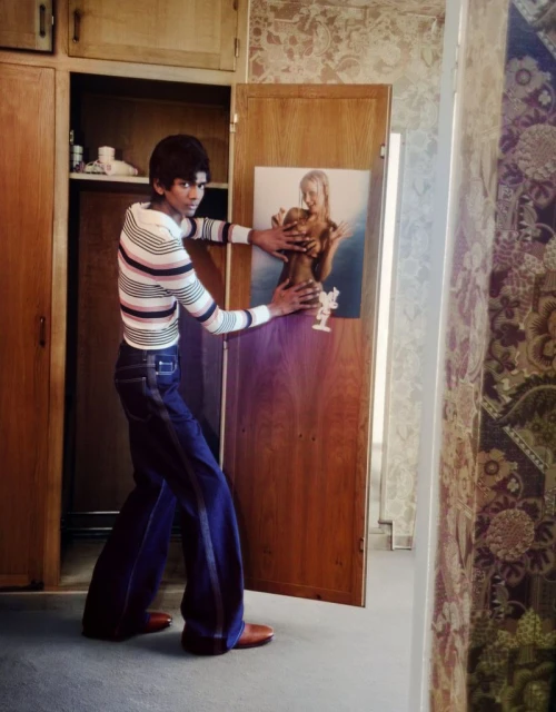 Man in striped shirt and bell-bottom jeans poses with a portrait of a woman attached to a wooden door in an indoor setting.