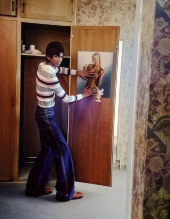 Man in striped shirt and bell-bottom jeans poses with a portrait of a woman attached to a wooden door in an indoor setting.
