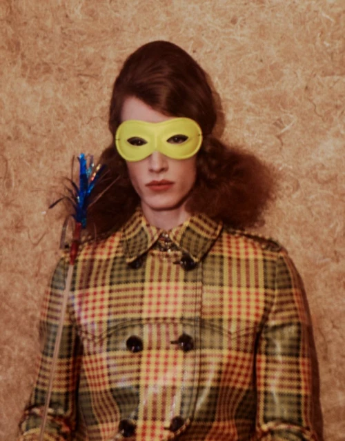 Woman wearing a plaid coat and a yellow mask holds a decorative stick in a studio shot with a textured beige background.