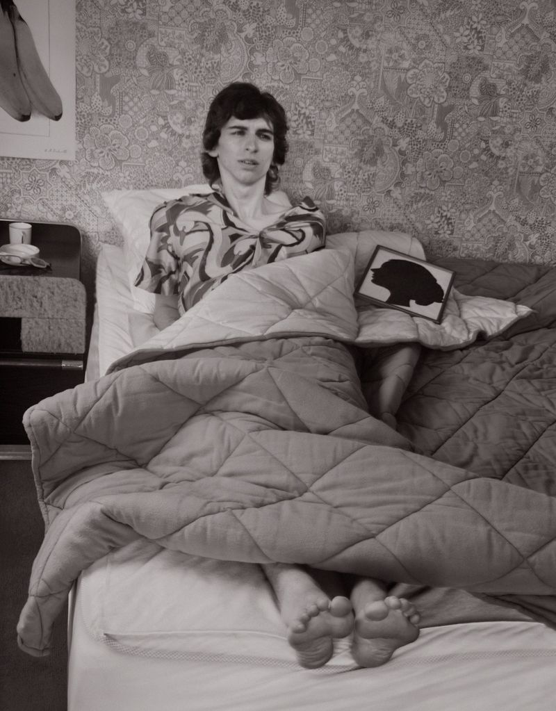 Monochrome photograph of a person in patterned shirt sitting in bed with a blanket, featuring floral wallpaper and a banana poster.
